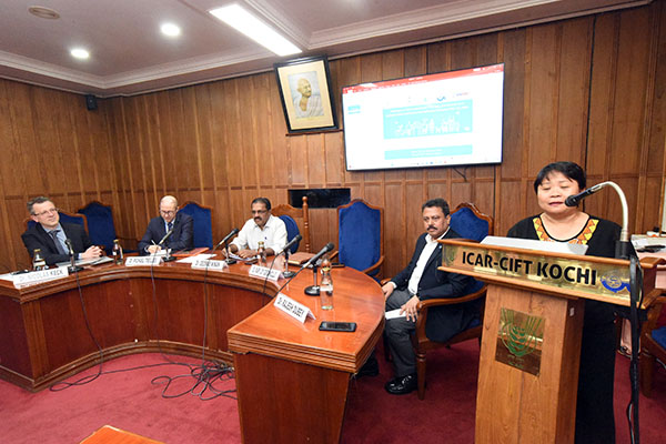Woman behind a podium while four men watching on behind a table.