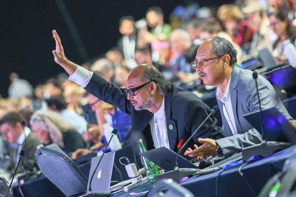 Two men sitting one raising his right hand